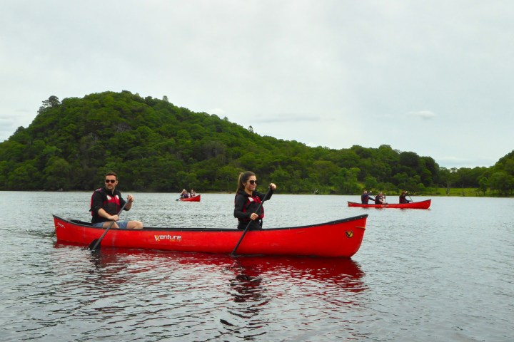 People in red canoes paddling on a lake with a green hill in the background.
