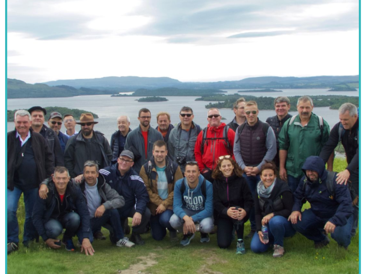 A group of people posing outdoors on grass with a lake and hills in the background.