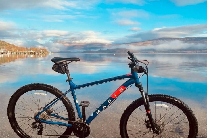 Blue bike on pebble shore against scenic lake and mountain view under cloudy sky.