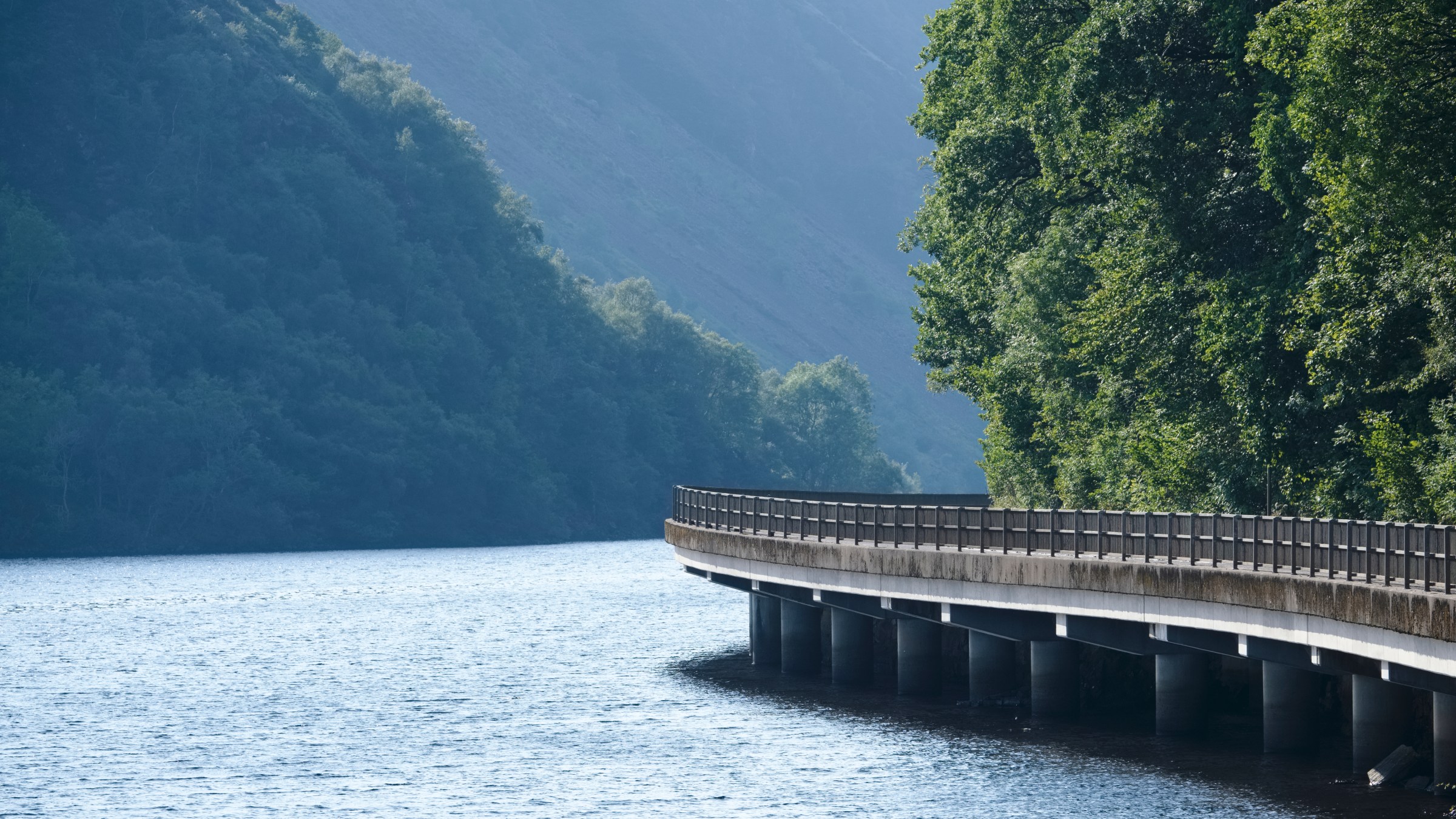 Curved bridge over a calm river with forested hills on both sides.
