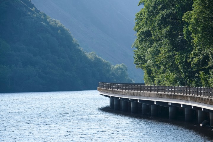Curved bridge over a calm river with forested hills on both sides.