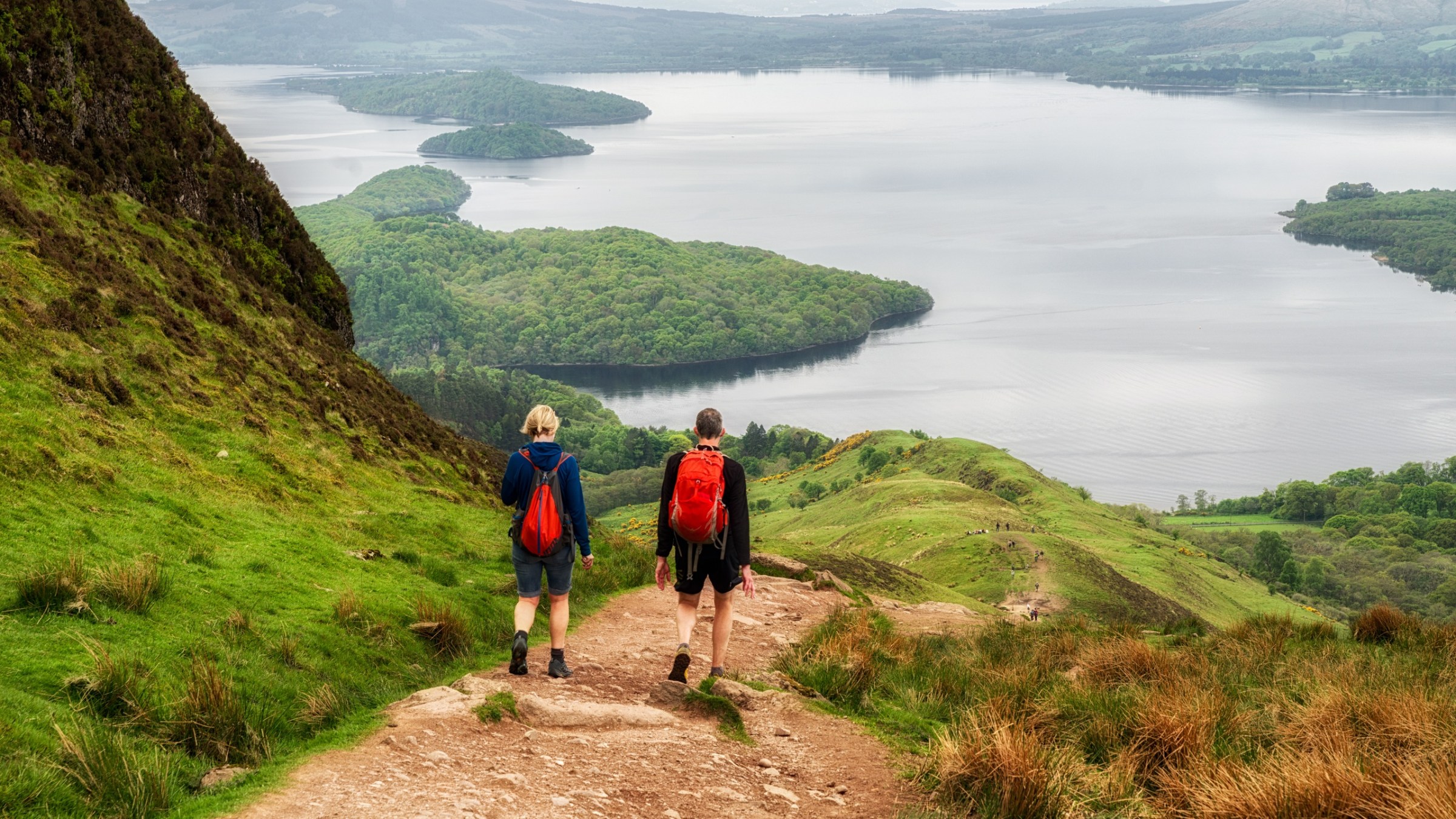 Two hikers with backpacks walking down a hill towards a lake view.