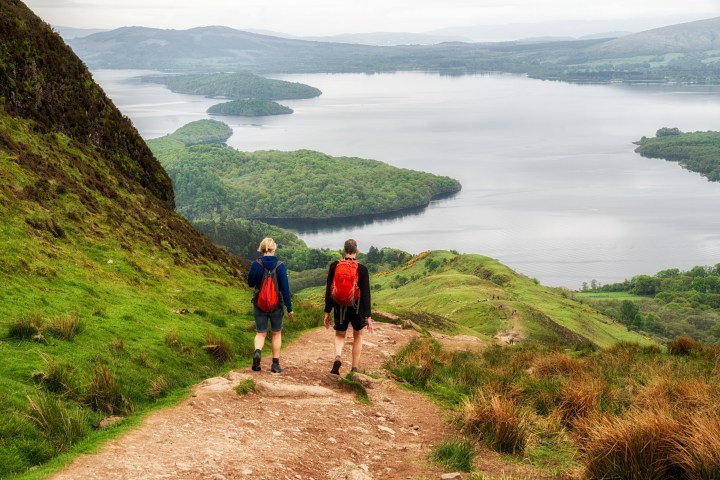 Two hikers with backpacks walking down a hill towards a lake view.