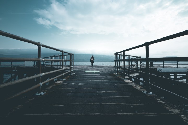 Person walking on pier towards water under cloudy sky.