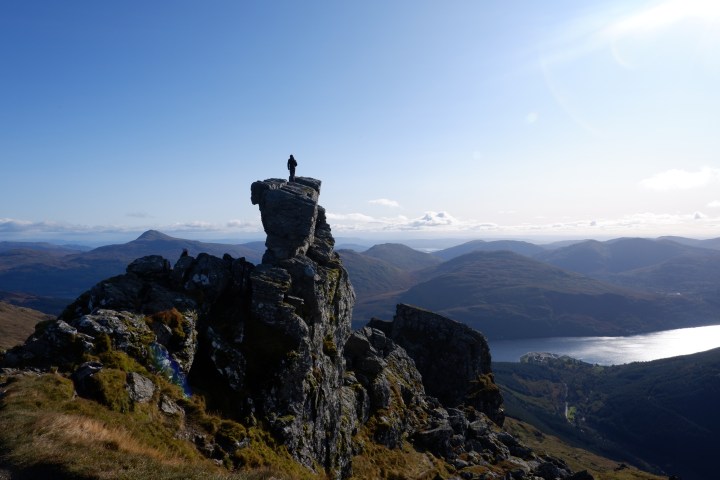 Person standing on a rocky peak overlooking a vast mountain landscape and lake.