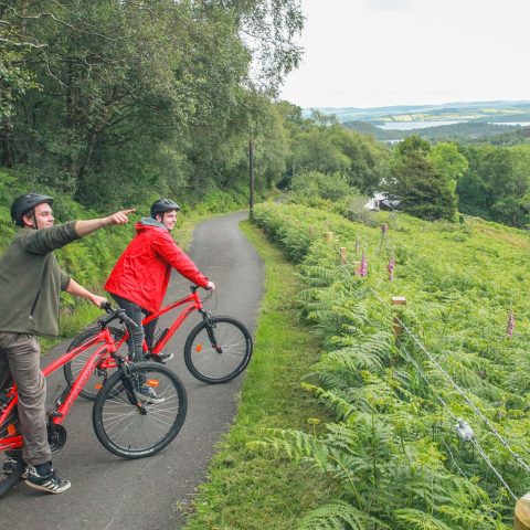 Two cyclists on a path in a lush, green landscape, one pointing ahead.