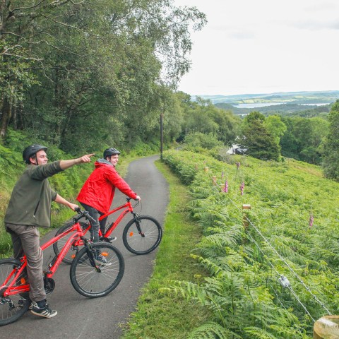 Two cyclists on a path in a lush, green landscape, one pointing ahead.