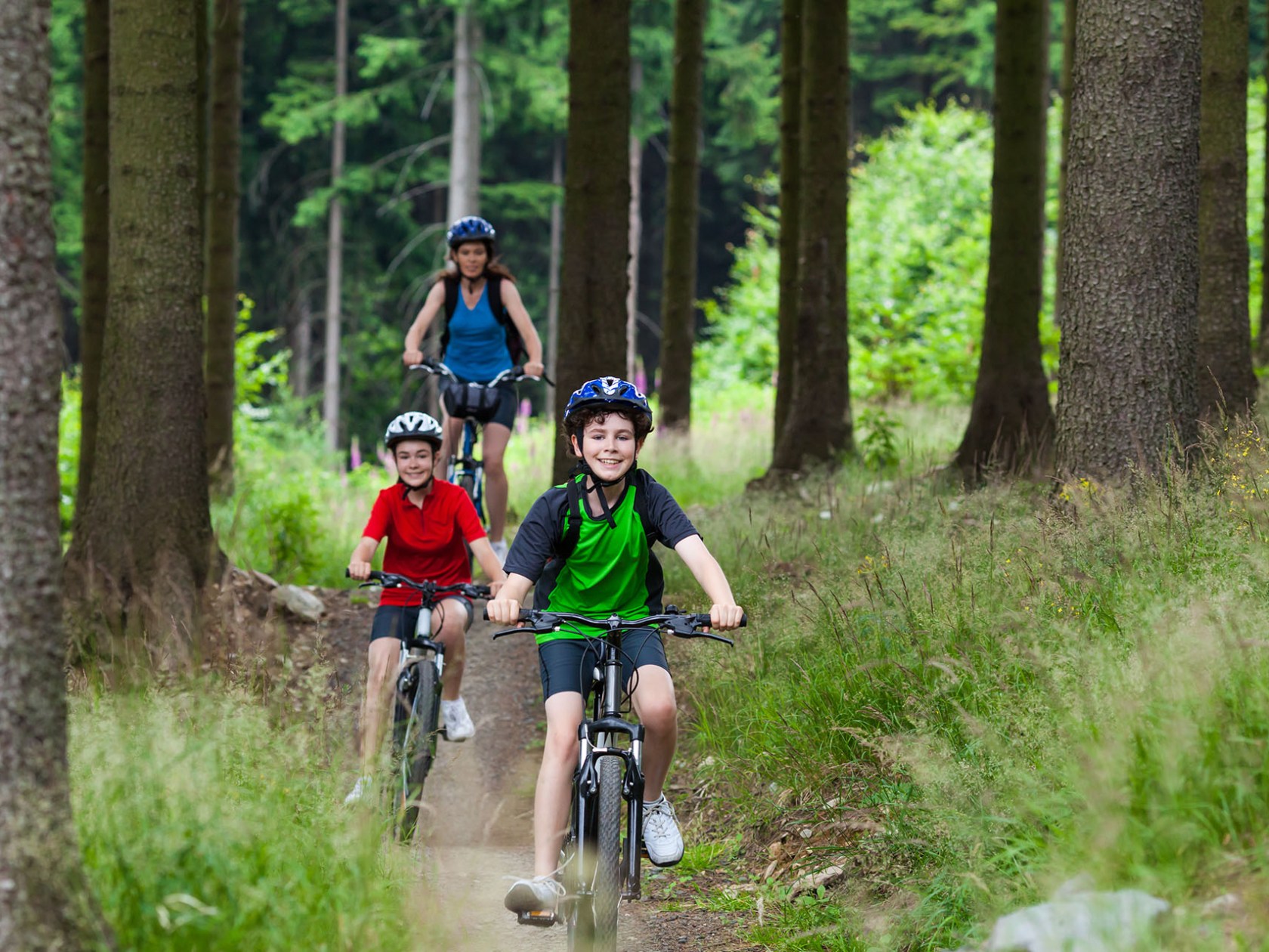 Three people biking on a forest trail, wearing helmets and casual clothes.