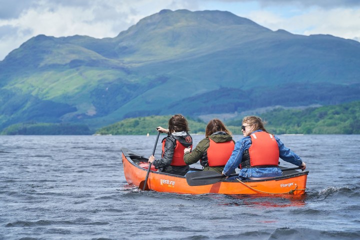 Three people in life vests canoeing on a lake with a mountain backdrop.