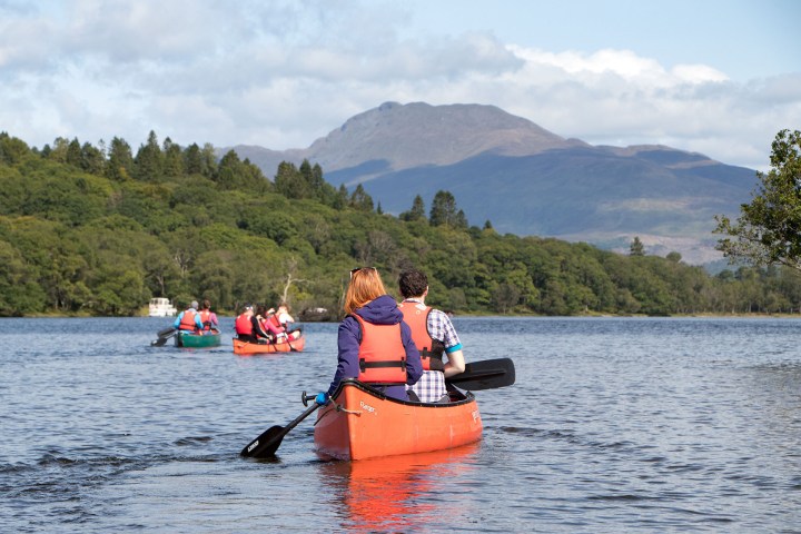 People canoeing on a lake with forested hills and mountains in the background.