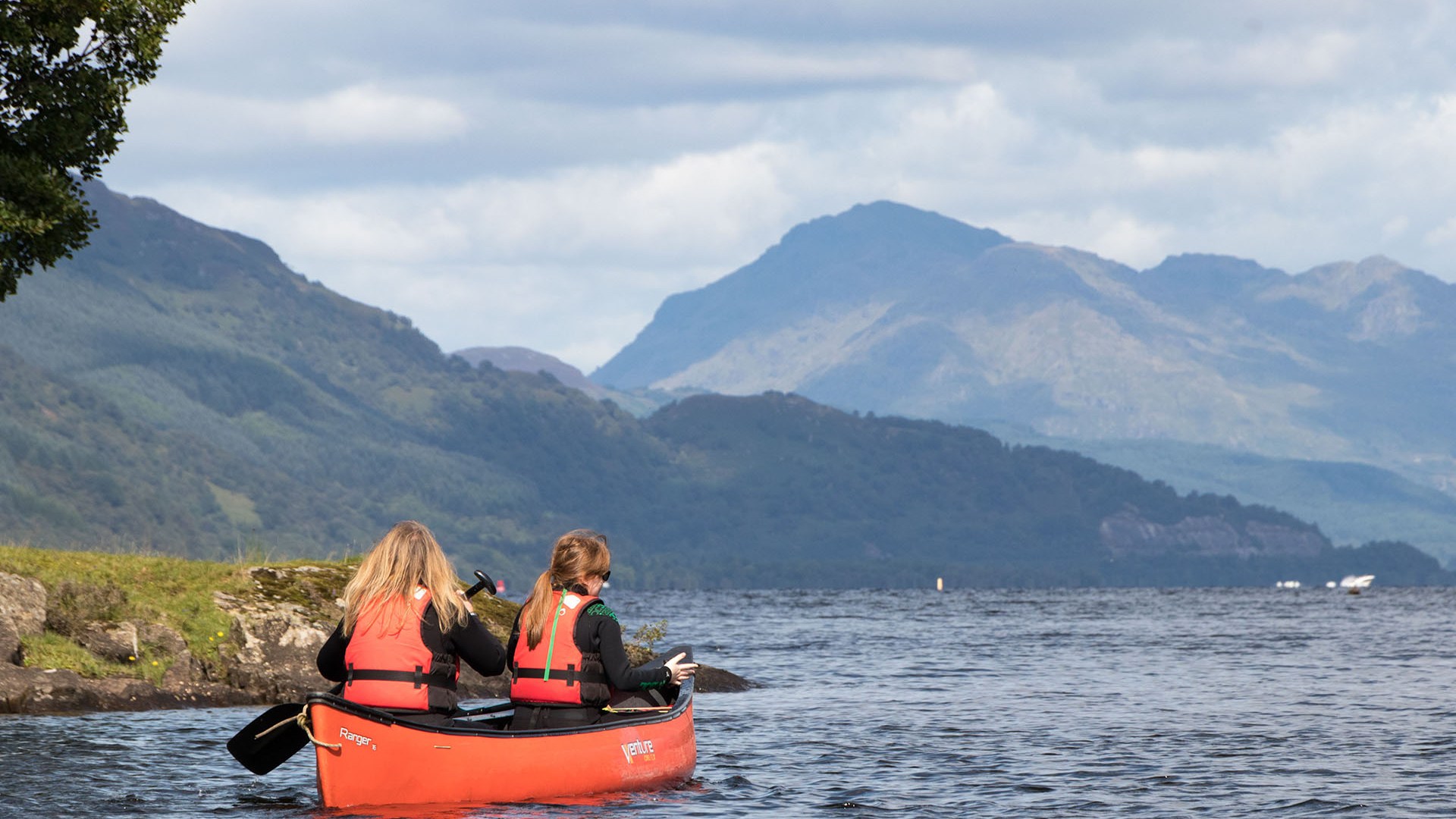 Two people in red life jackets canoeing on a lake with mountains in the background.