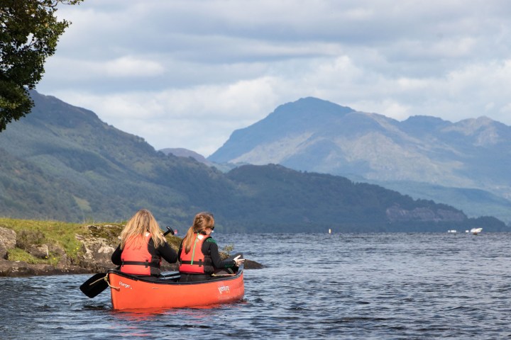 Two people in red life jackets canoeing on a lake with mountains in the background.