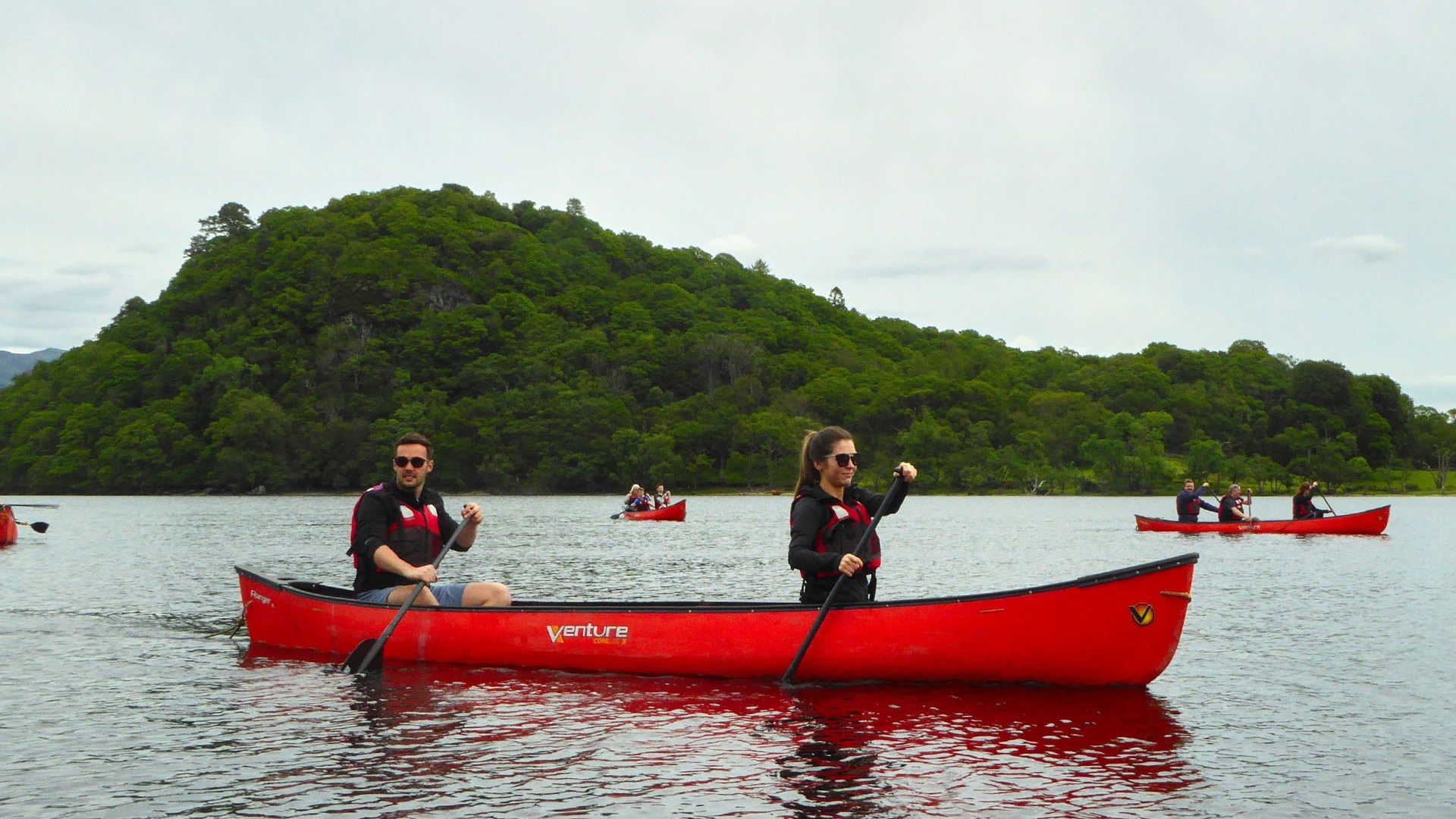 Two people in a red canoe paddling on a lake with a green hill in the background.
