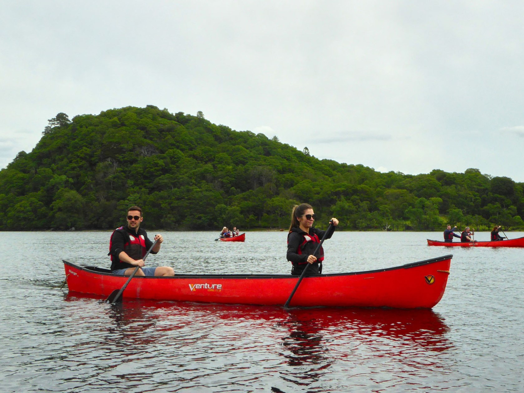 Two people in a red canoe paddling on a lake with a green hill in the background.