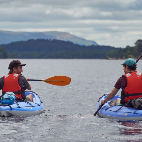 Two people kayaking on a lake with mountains and trees in the background.
