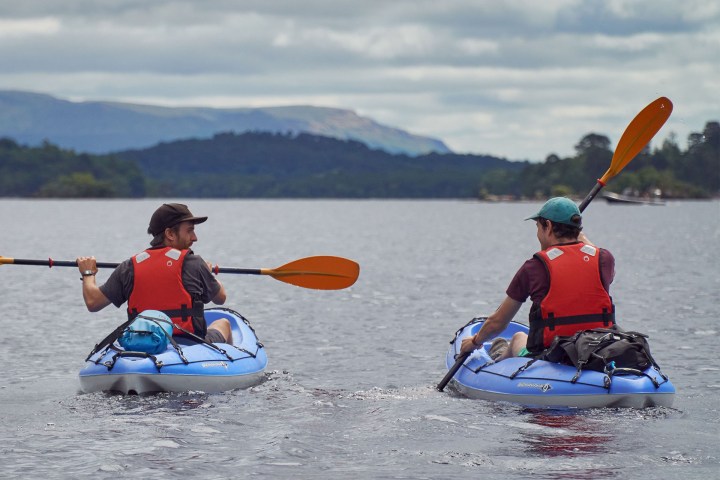 Two people kayaking on a lake with mountains and trees in the background.