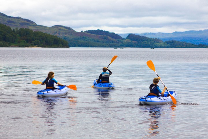 Three people kayaking on a calm lake with forested hills in the background.