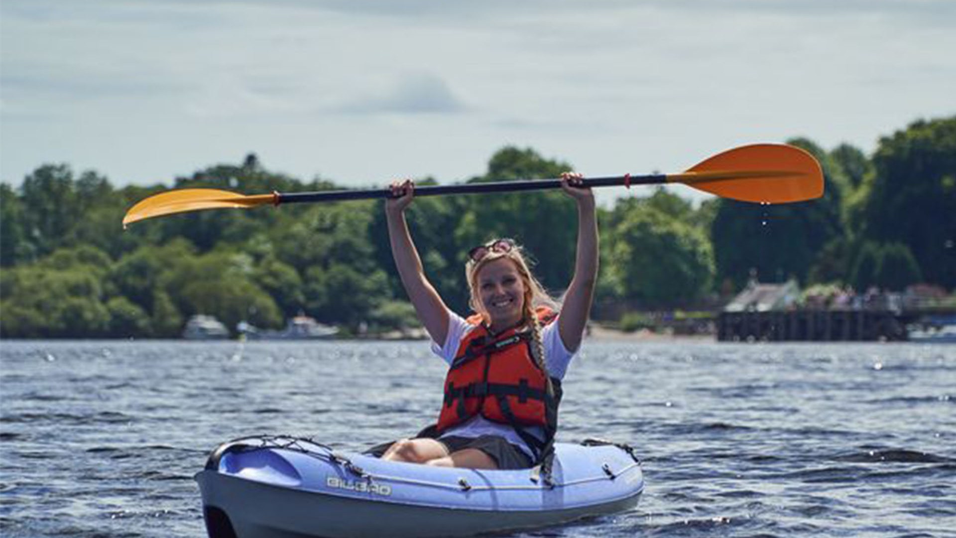 Person kayaking on a lake, holding a paddle overhead, wearing a red life jacket.
