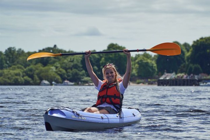 Person kayaking on a lake, holding a paddle overhead, wearing a red life jacket.