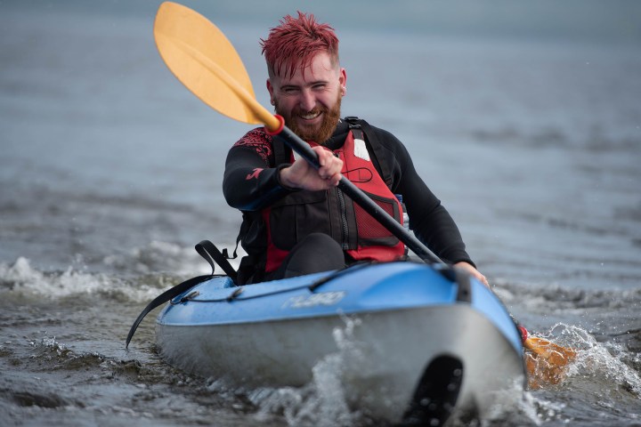 Person with red hair kayaking on water in a blue kayak with a yellow paddle.