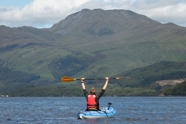 Person in kayak raises paddle triumphantly on a lake with mountains in the background.