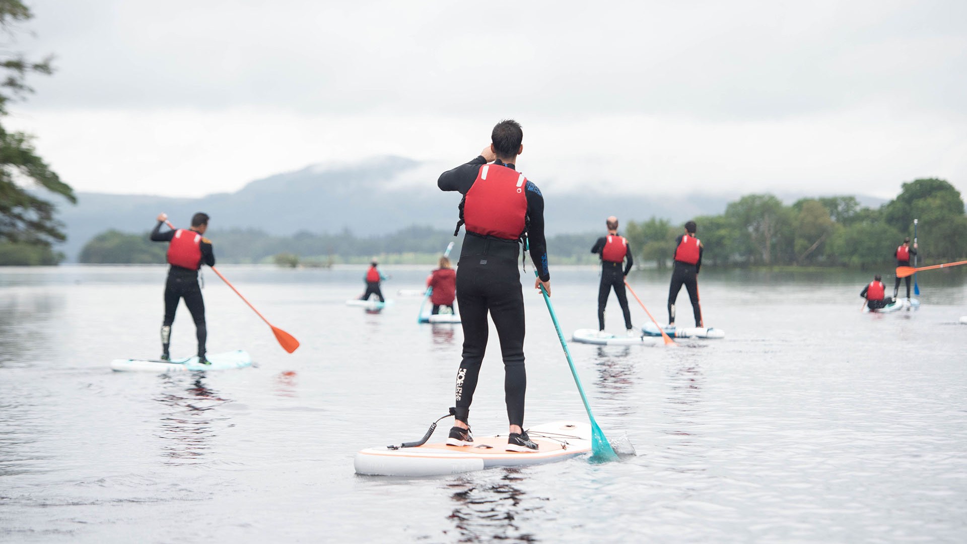 Group paddleboarding on a calm lake, wearing red life jackets and wetsuits.