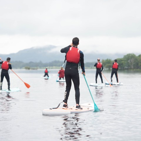 Group paddleboarding on a calm lake, wearing red life jackets and wetsuits.