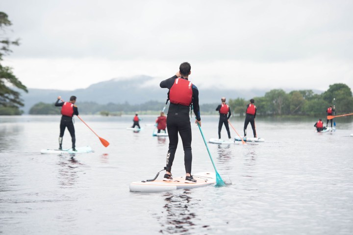 Group paddleboarding on a calm lake, wearing red life jackets and wetsuits.