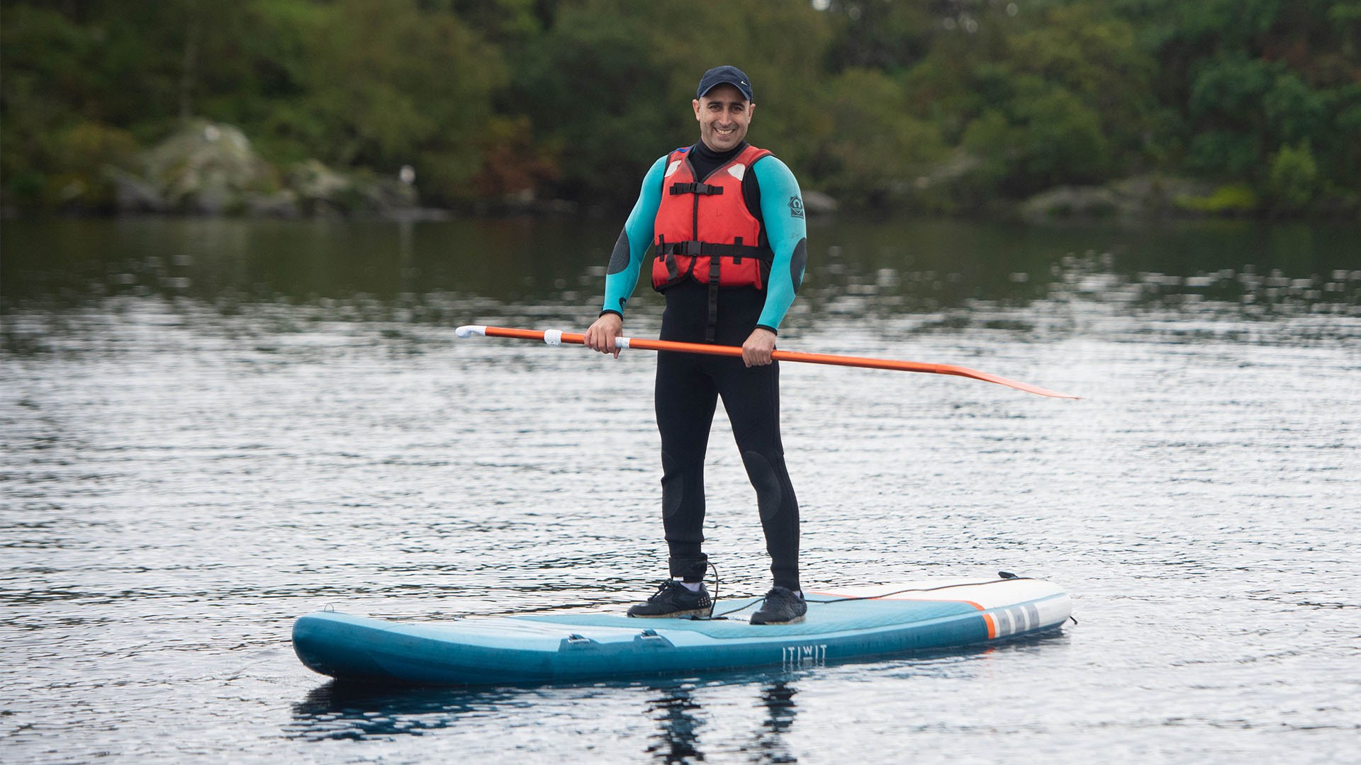 Person in wetsuit and life jacket paddleboarding on a lake with trees in the background.