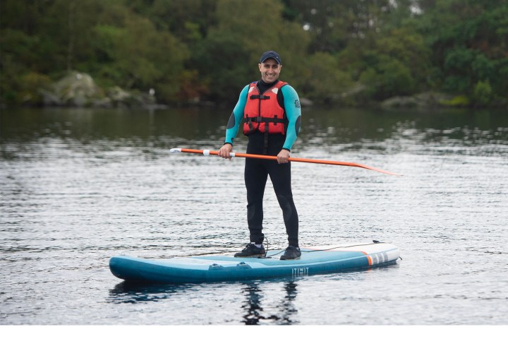 Person in wetsuit and life jacket paddleboarding on a lake with trees in the background.