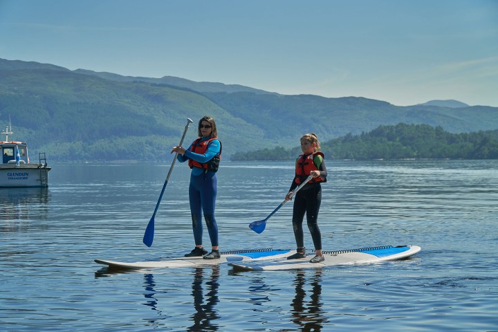 Two people paddleboarding on a calm lake with life vests; scenic hills in the background.