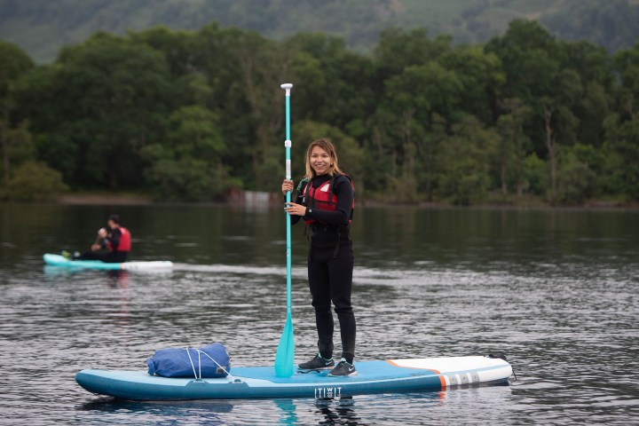 Person paddleboarding on a lake, wearing a wetsuit and life vest, with trees in the background.