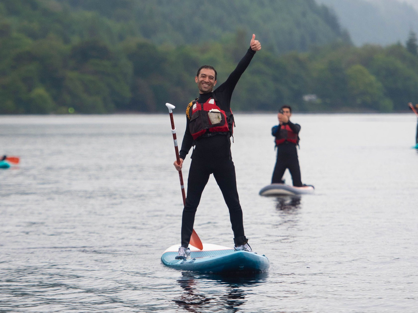 Person paddleboarding on a lake gives a thumbs-up, with others in the background.