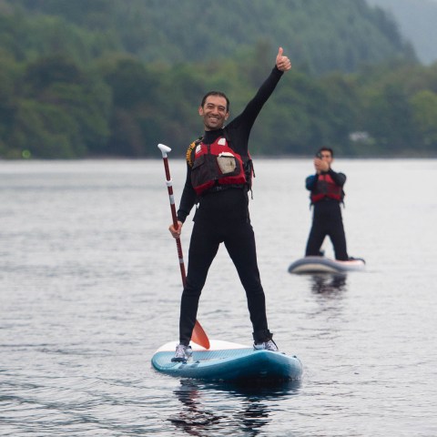 Person paddleboarding on a lake gives a thumbs-up, with others in the background.