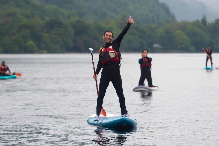 Person paddleboarding on a lake gives a thumbs-up, with others in the background.