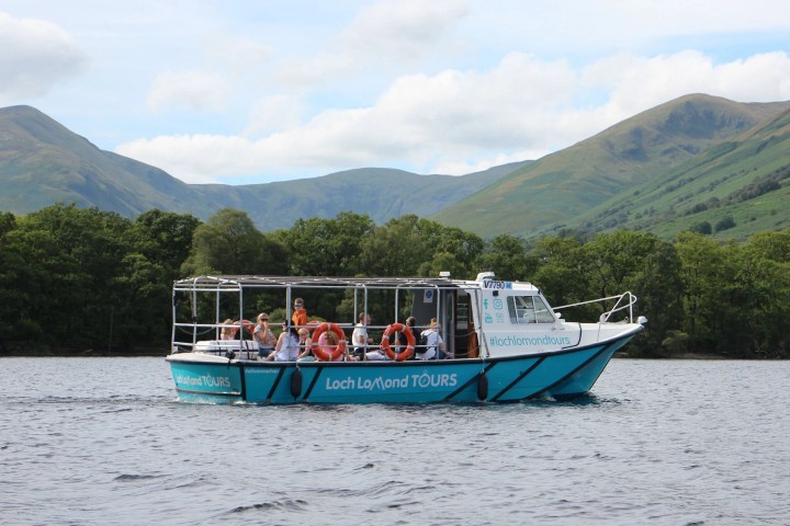 Tour boat on a lake with passengers, surrounded by green hills and trees.