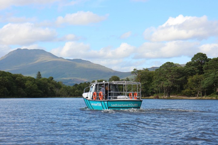 Tour boat on Loch Lomond with mountains and trees in the background, under a blue sky with clouds.