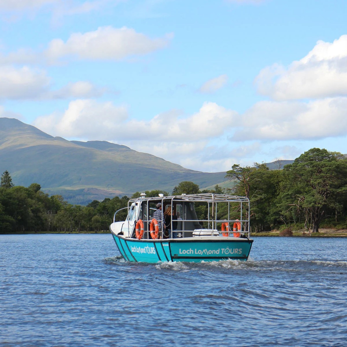Tour boat on Loch Lomond with mountains and trees in the background, under a blue sky with clouds.
