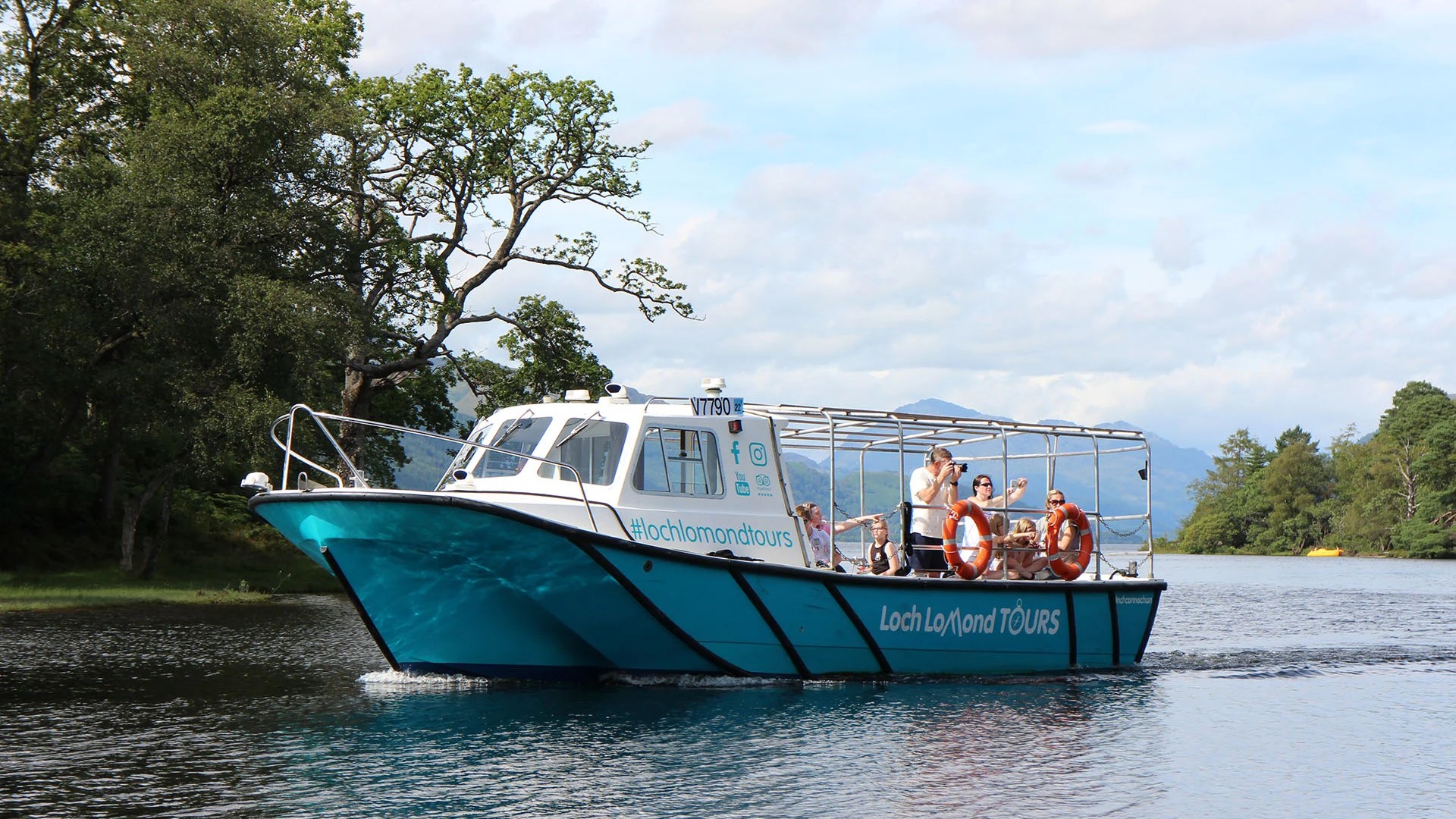 Tour boat on a lake with trees and mountains in the background under a cloudy sky.