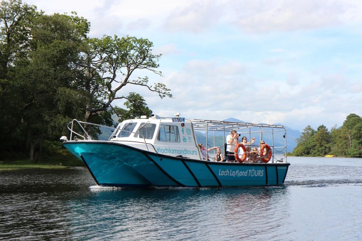 Tour boat on a lake with trees and mountains in the background under a cloudy sky.