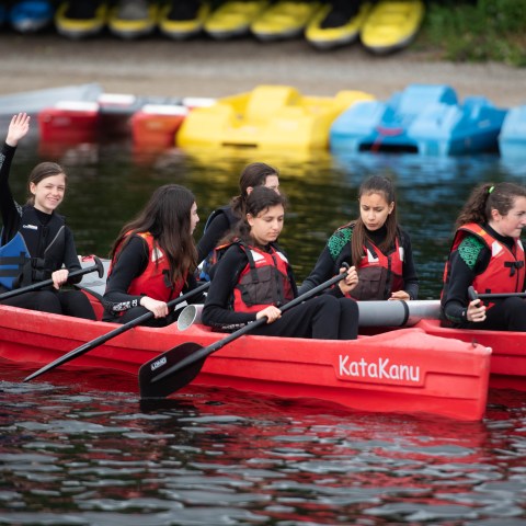 Group of girls in life jackets paddling a red kayak on a lake, one girl waves.