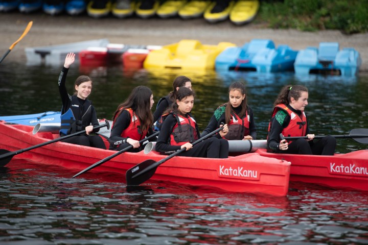Group of girls in life jackets paddling a red kayak on a lake, one girl waves.
