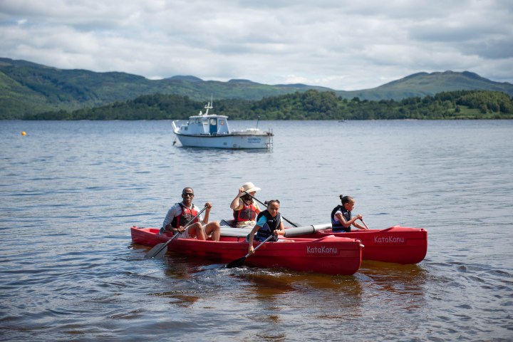 Two red canoes with people paddling on a lake, boat and green hills in the background.
