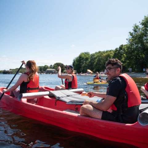 Group kayaking on a lake, wearing red life vests, with trees and clear sky in the background.