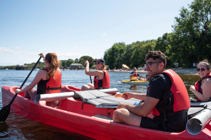 Group kayaking on a lake, wearing red life vests, with trees and clear sky in the background.